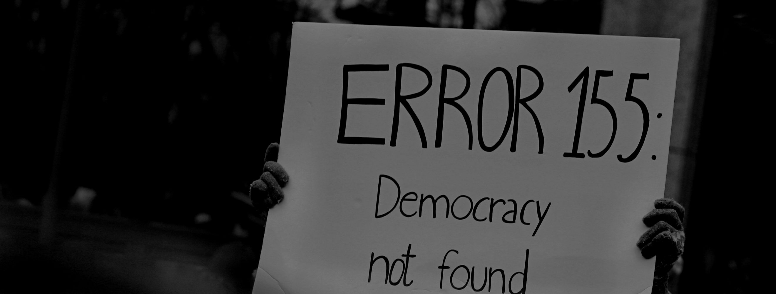 A person holding a protest sign, covering the persons face, with the text: Error 155: Democracy not found. The picture is black and white.