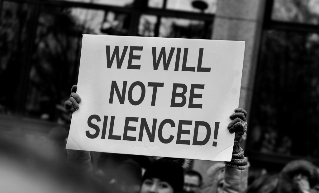 Protestskylt med texten ”WE WILL NOT BE SILENCED!” hålls upp över en folkmassa vid en demonstration i stadsmiljö.