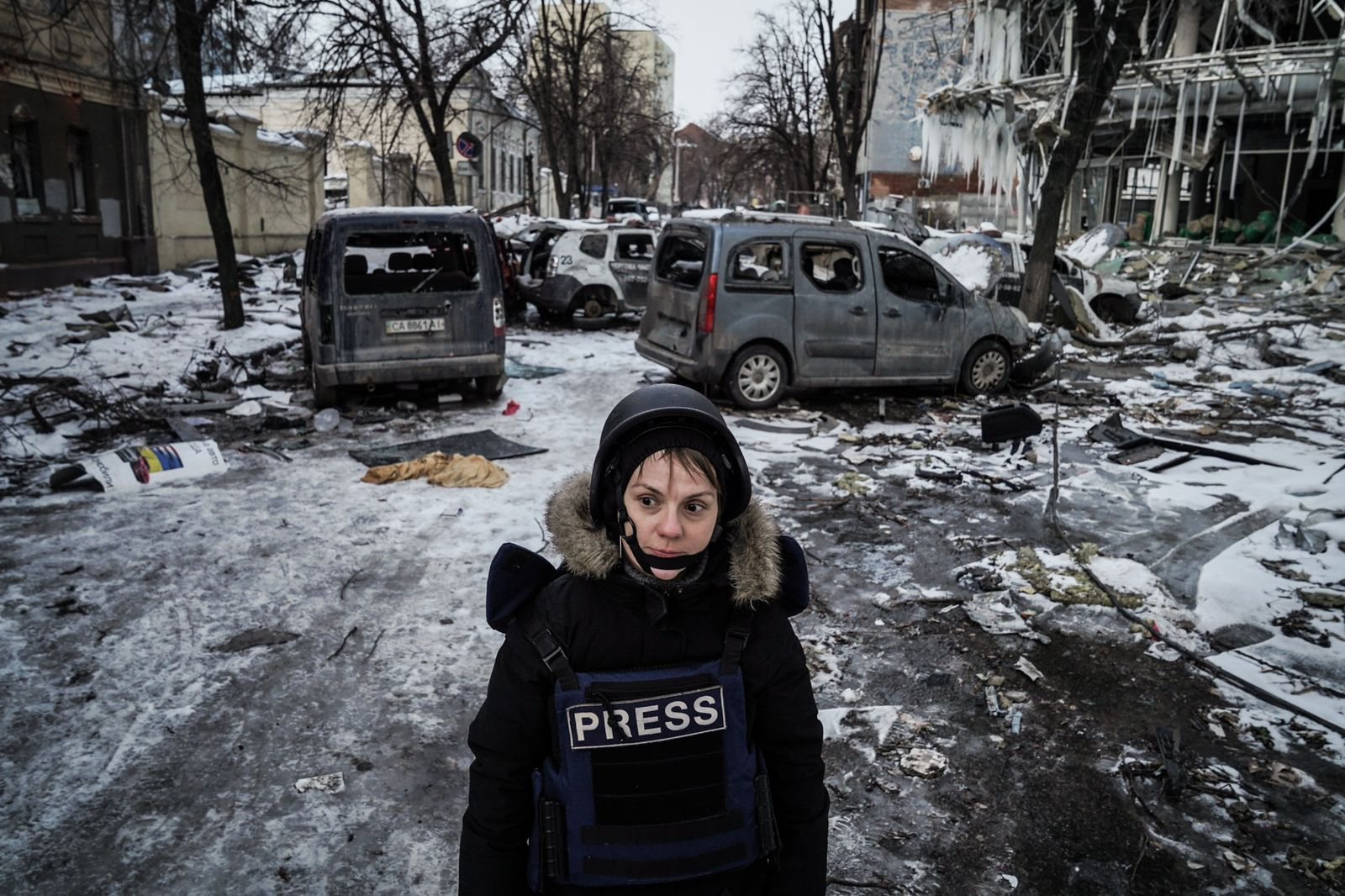Nataliya in helmet and vest with the text "press". Behind here bombed cars and buildings.
