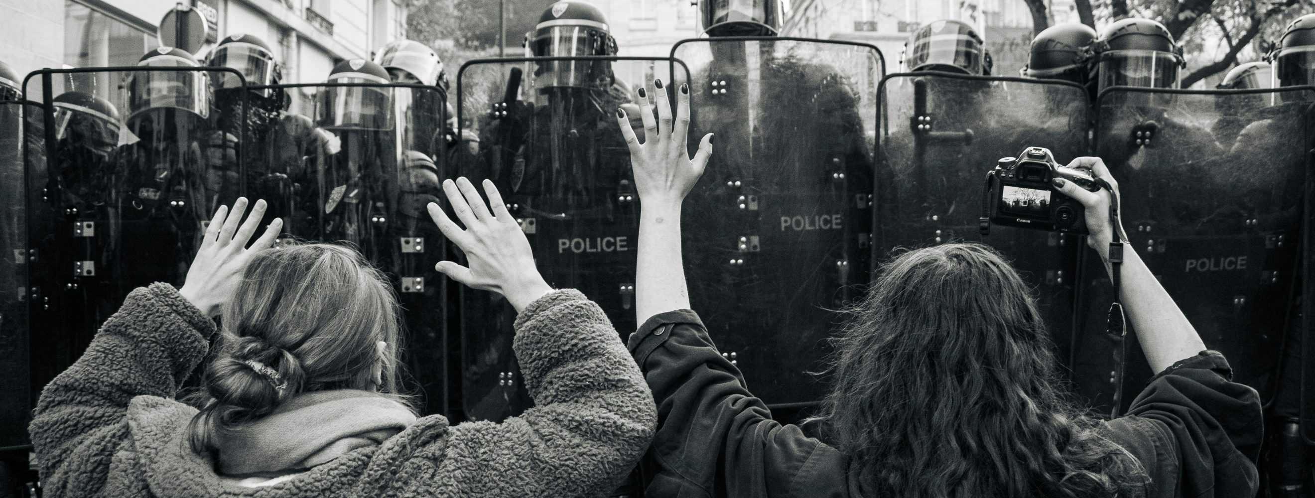 Two women are standing in front of a line of police officer in heavy gear during a protest.
