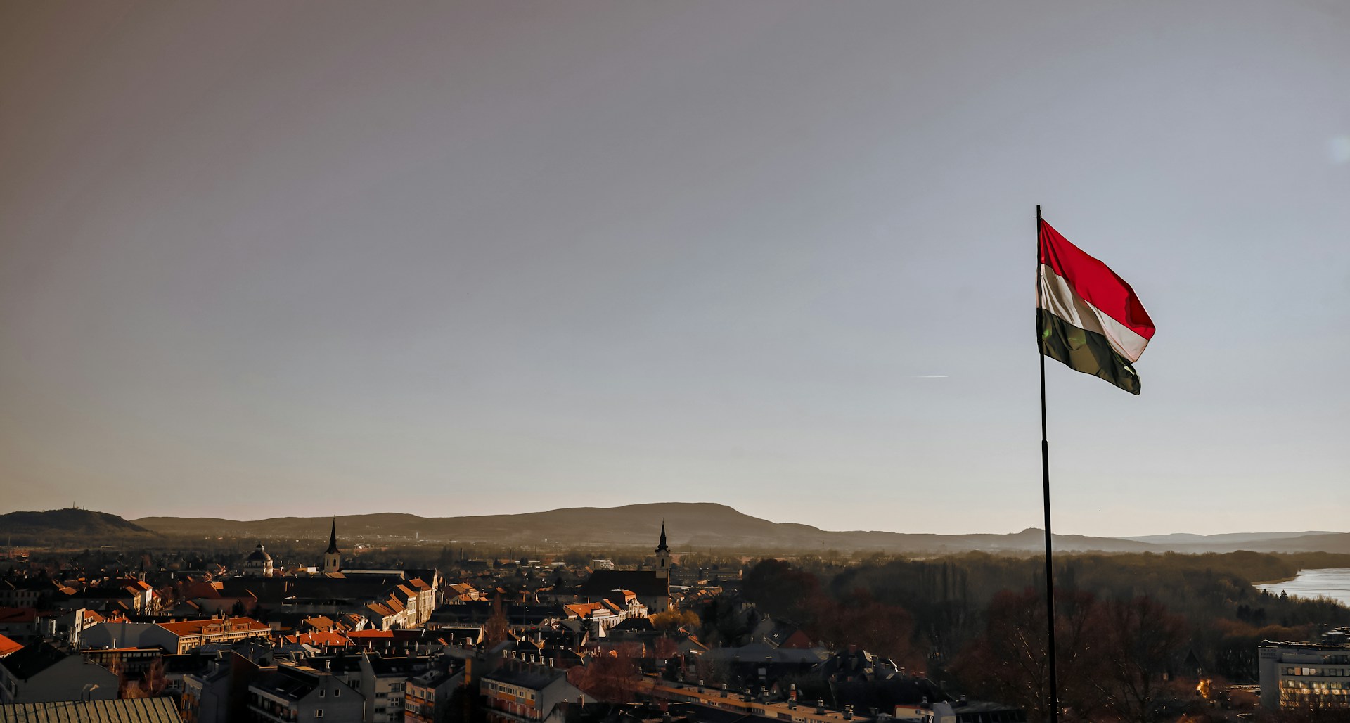 Hungarian flag blowing in the wind, against a backdrop of rolling hills and a town, around sunset