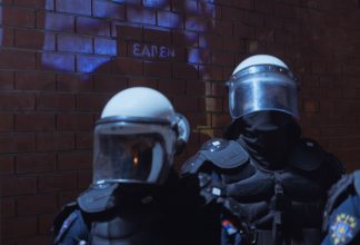 Police officers stand in front of a brick wall with the word 'Exit' faintly visible behind them.