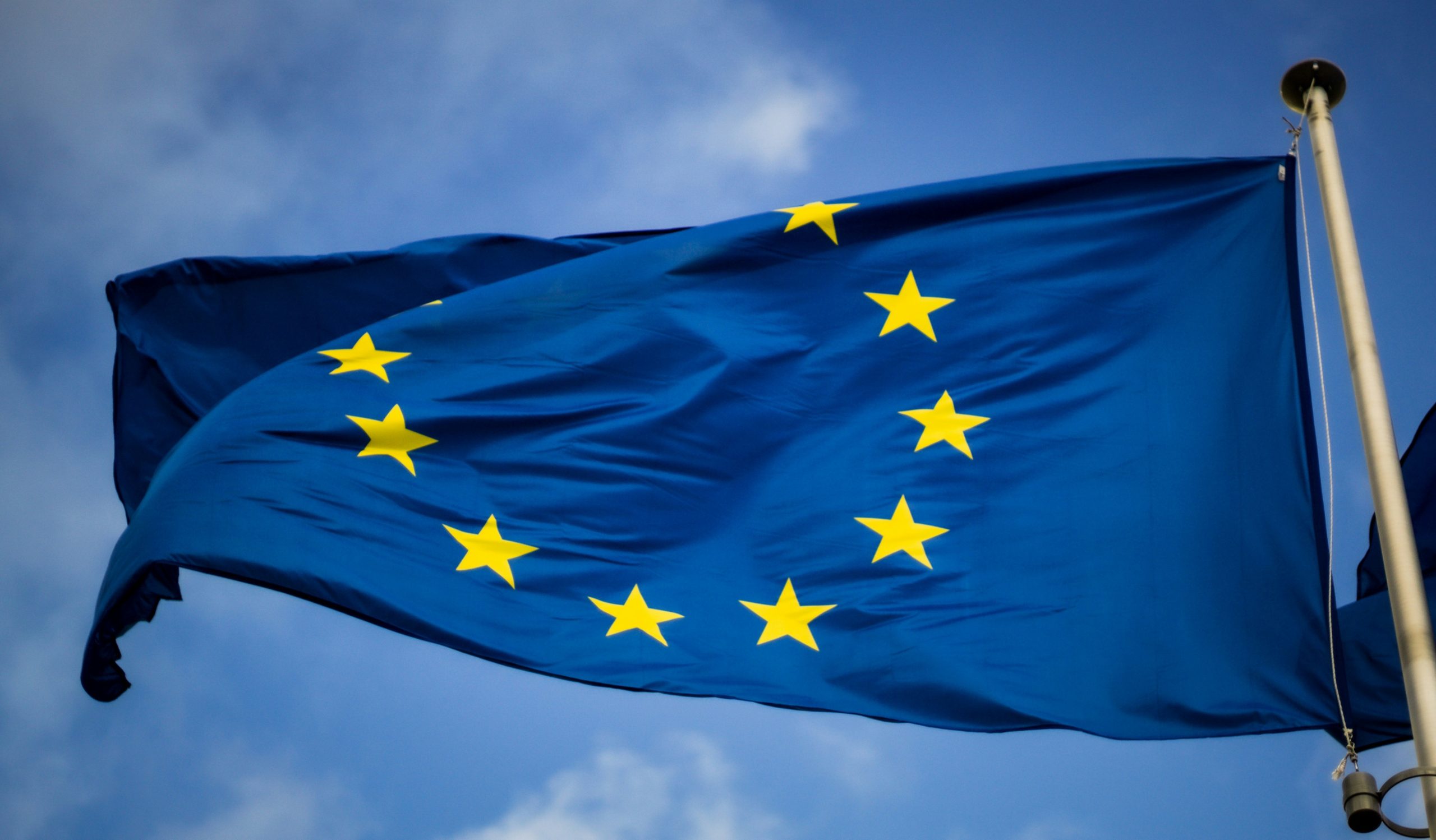 The EU-flag is blowing in the wind in the setting of a blue sky. 