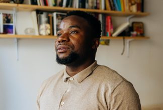 Dieu-Merci har ett neutralt ansiktsuttryck medan han tittar bort från kameran i en lägenhet med beige väggar.