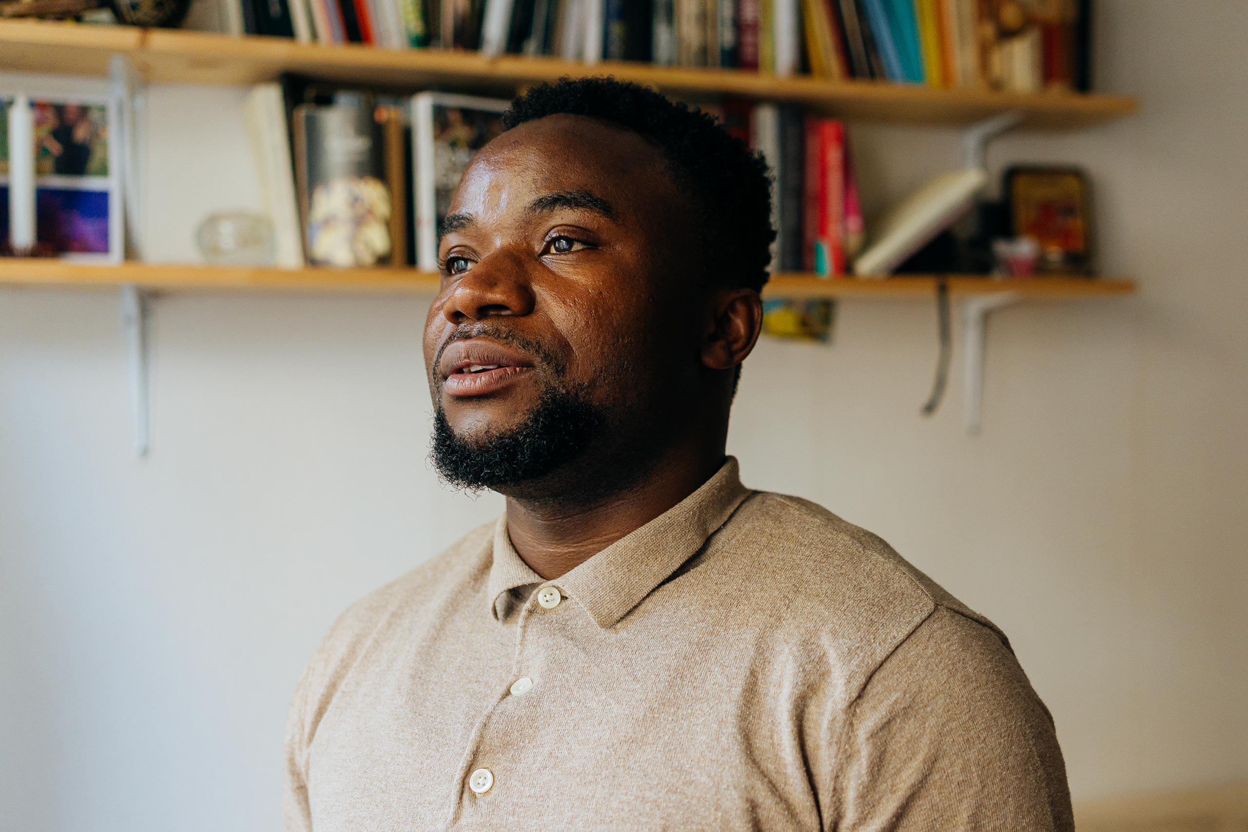 Dieu Merci har ett neutralt ansiktsuttryck medan han tittar bort från kameran i en lägenhet med beige väggar.