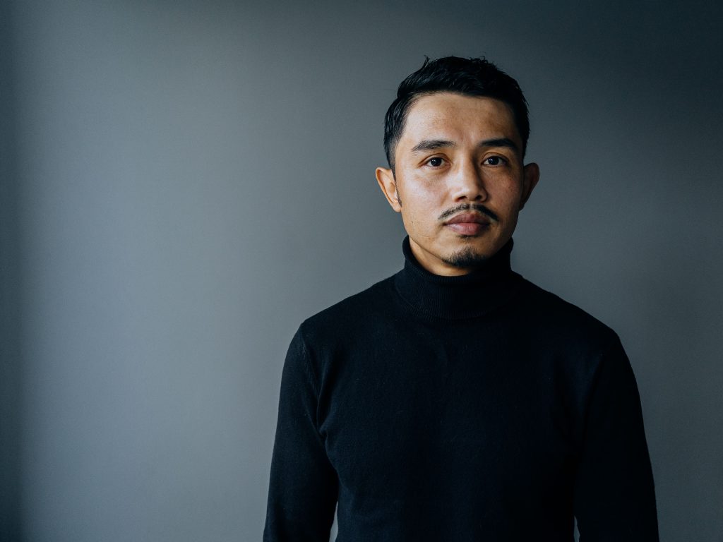 Ali Yusefi stands against a dark background, looking into the camera with a neutral expression.