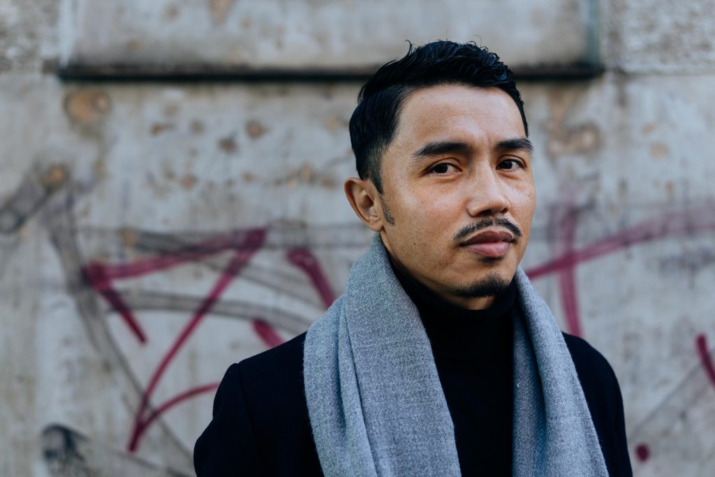 Ali Yusefi stands against a dark background, looking into the camera with a neutral expression.