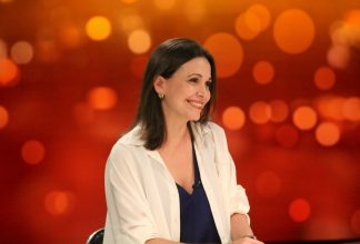 Maria Corina Machado from Venezuela, wearing a white shirt and smiling in front of a reddish orange backdrop