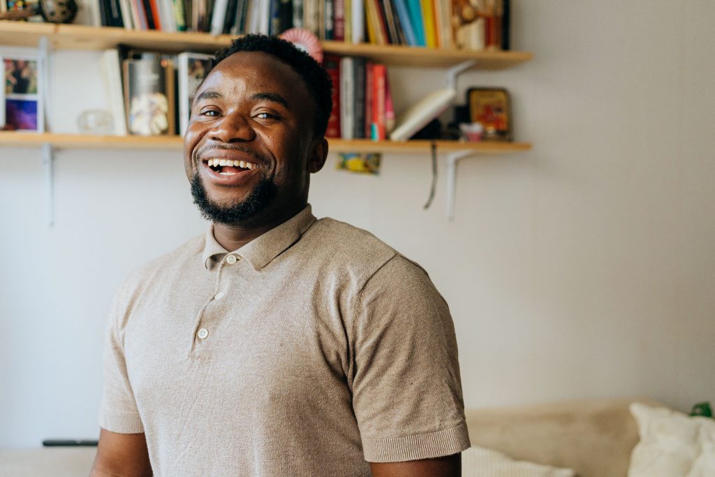 Dieu Merci skrattar och tittar in i kameran i en lägenhet med beige väggar.