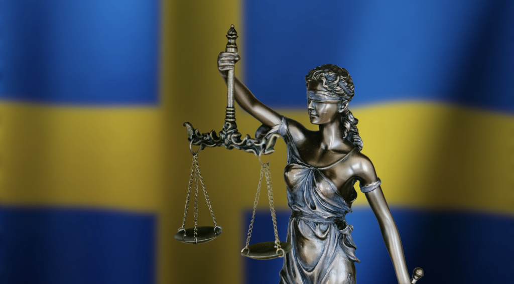Justitia‑statyn som håller vågskålar framför svenska flaggan – symbol för rättssäkerhet i Sverige.