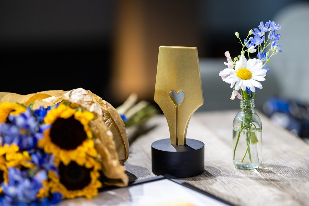 The Civil Rights Defenders of the Year Award standing on a table. In the foreground, a bouquet of sunflowers and blue flowers wrapped in brown paper. To the right, a small glass bottle with a daisy and blue wildflowers.