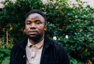 Dieu-Merci står framför ett buskage utomhus i Stockholm och tittar in i kameran.